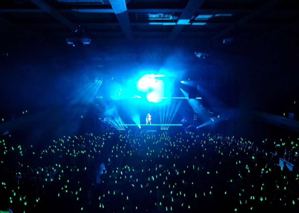 A picture of Miku Expo 2026, there are thousands of green light sticks and a bright blue screen with Hatsune Miku in the distance.