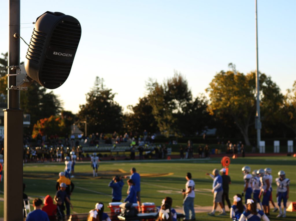Zoomed in photo of a high-school football field. A pole with a black speaker attached on the very left hand side, in focus. Football players and other team members on the field, out of focus.