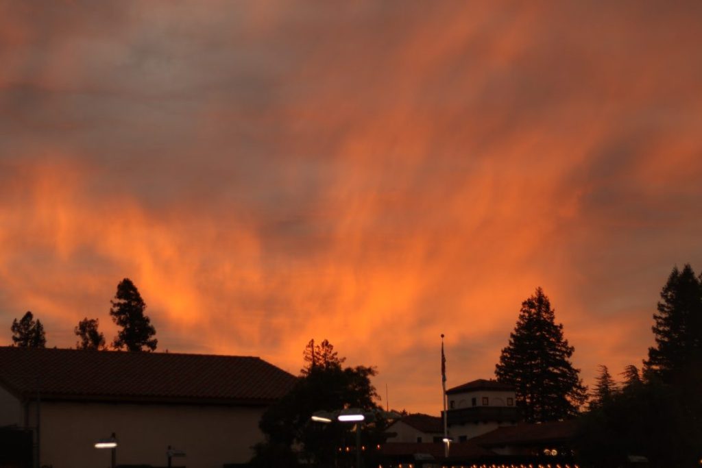 Clouds glowing from a beautiful orange-rich sunset with shadowed houses and trees in the foreground.