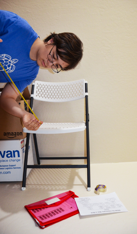 A scene showing a chair, stacked boxes, and clutter on the floor, with a human subject holding a measuring tape posing as if gravity direction was sideways rather than down.