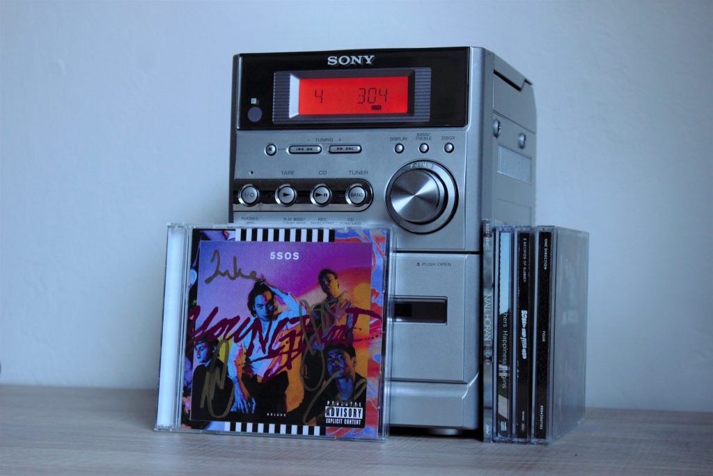 A photo of CD's displayed against a Sony CD player. A signed CD of the album "Youngblood" by 5 Seconds of Summer is leaning against the front of the CD player. There are four other CD's leaning against the side of the CD player, only showing their spines.