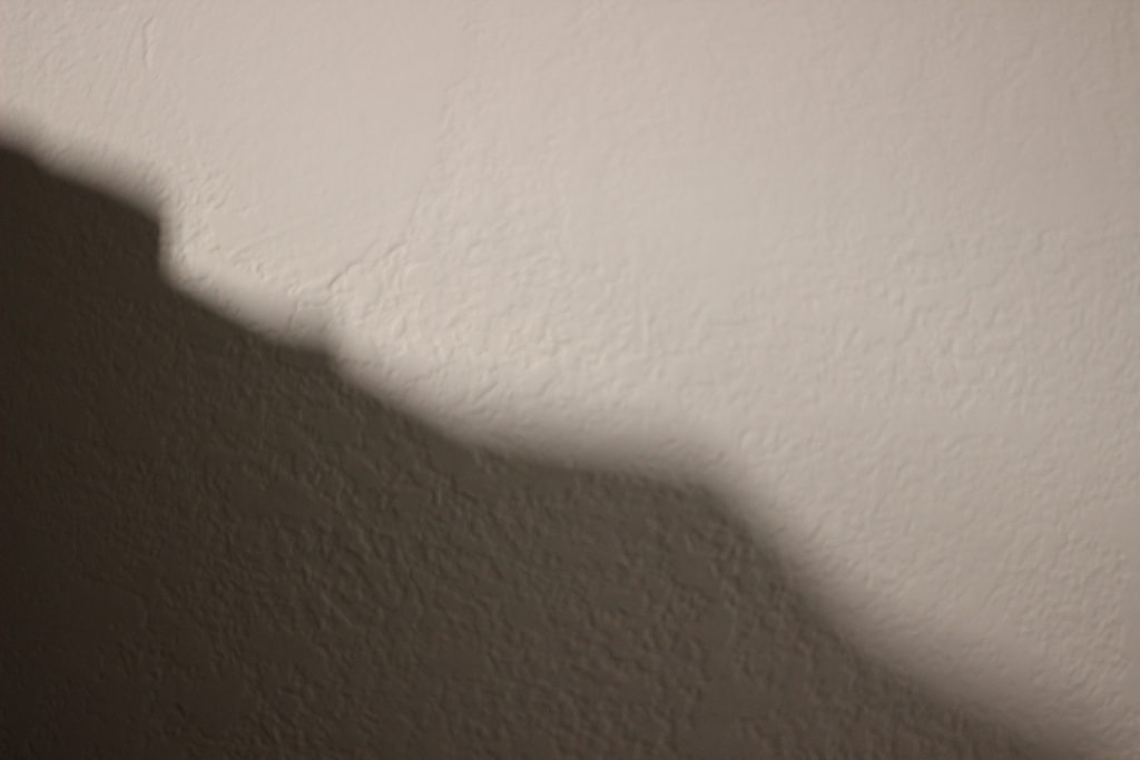 A shadow cast on a white wall. The shadow looks like a hill going downward diagonally starting in the top left corner.