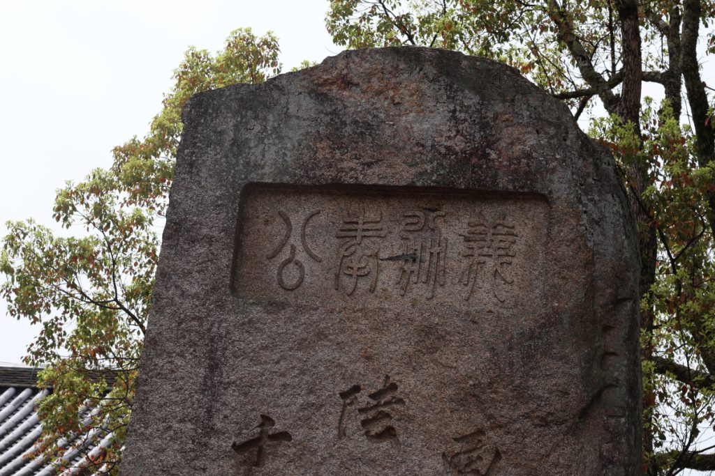 photo of an old stone with Japanese characters carved into it. The stone is worn down and shows very many colors within it. 
