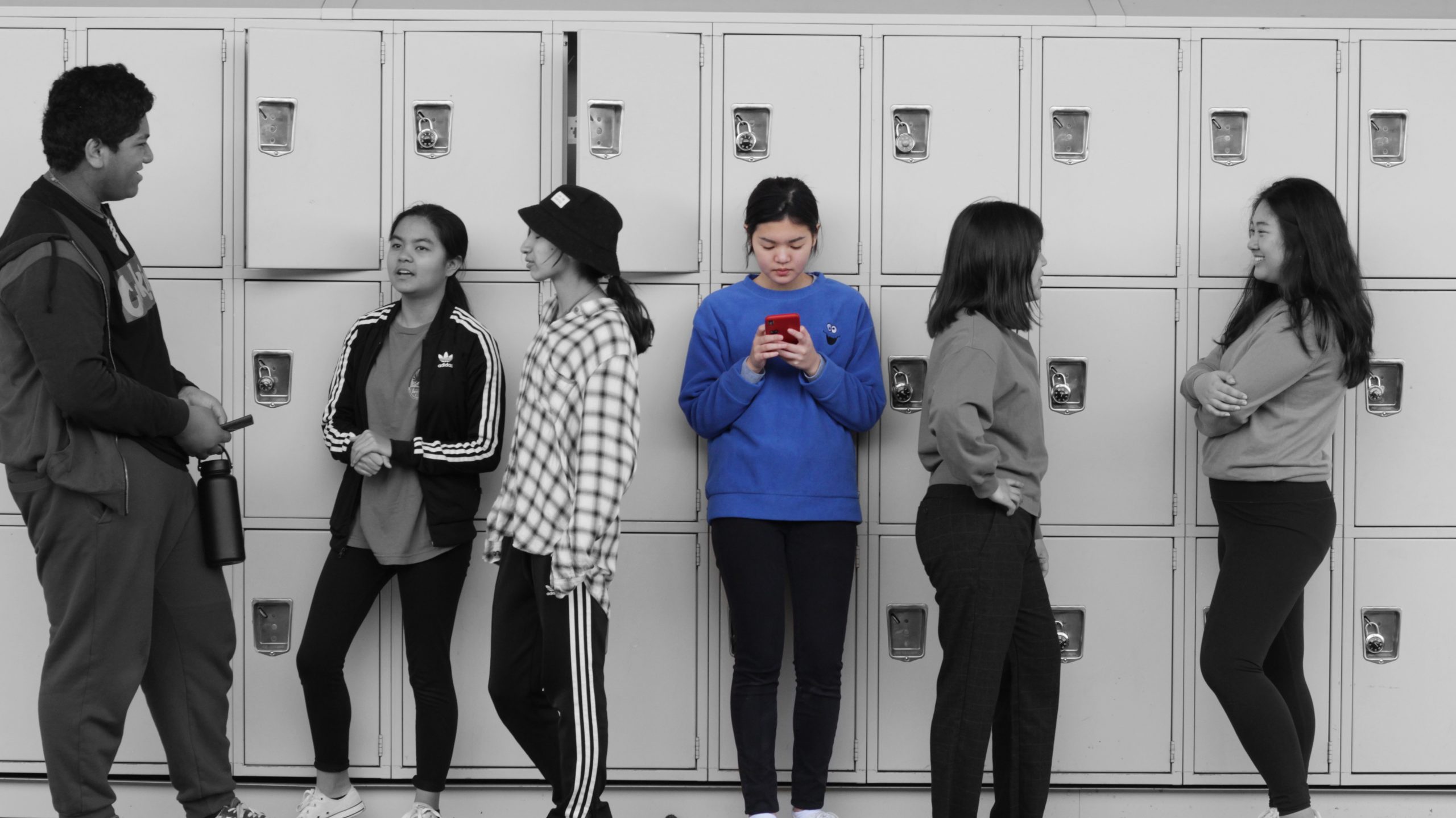 Photo of an antisocial girl on her phone in the center, surrounded by socializing groups of teenagers in black and white.