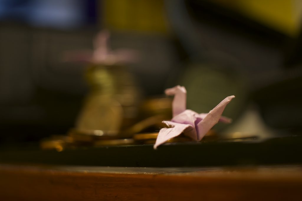 Paper crane with a ripped wing on the ground watches another crane on top of a tower of coins.