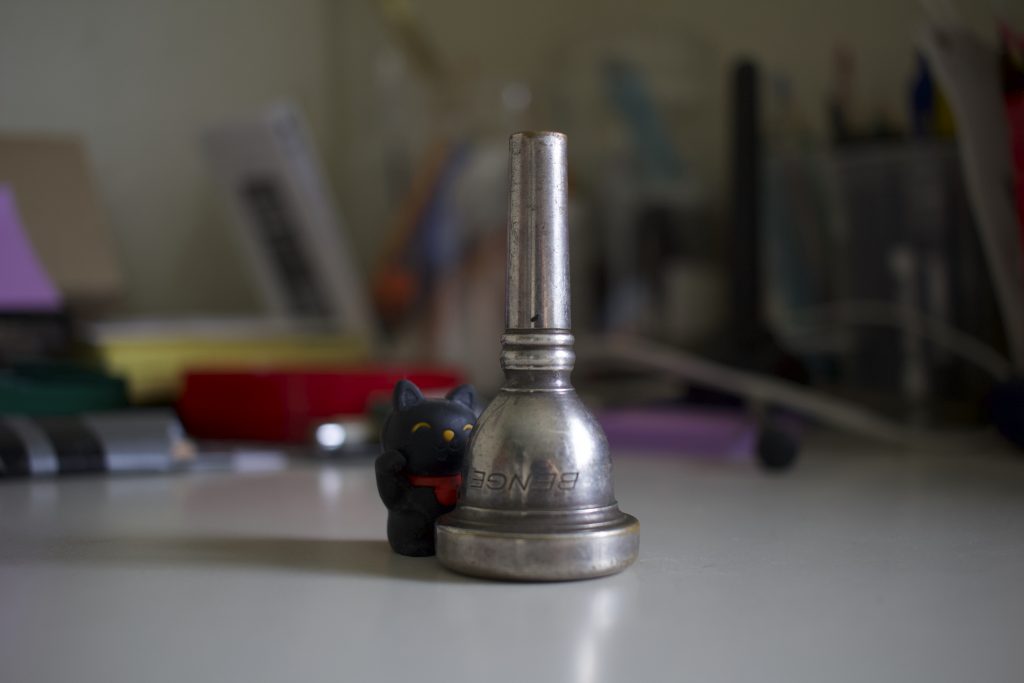 Black Cat Eraser behind a metal trombone mouthpiece, set in a clearing of a messy desk.