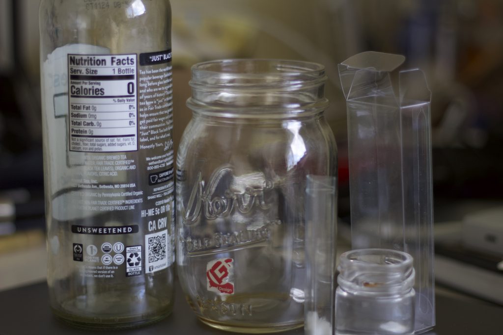 Assortment of clear wastes on top of a metal surface- a glass bottle, two glass jars, and plastic containers.