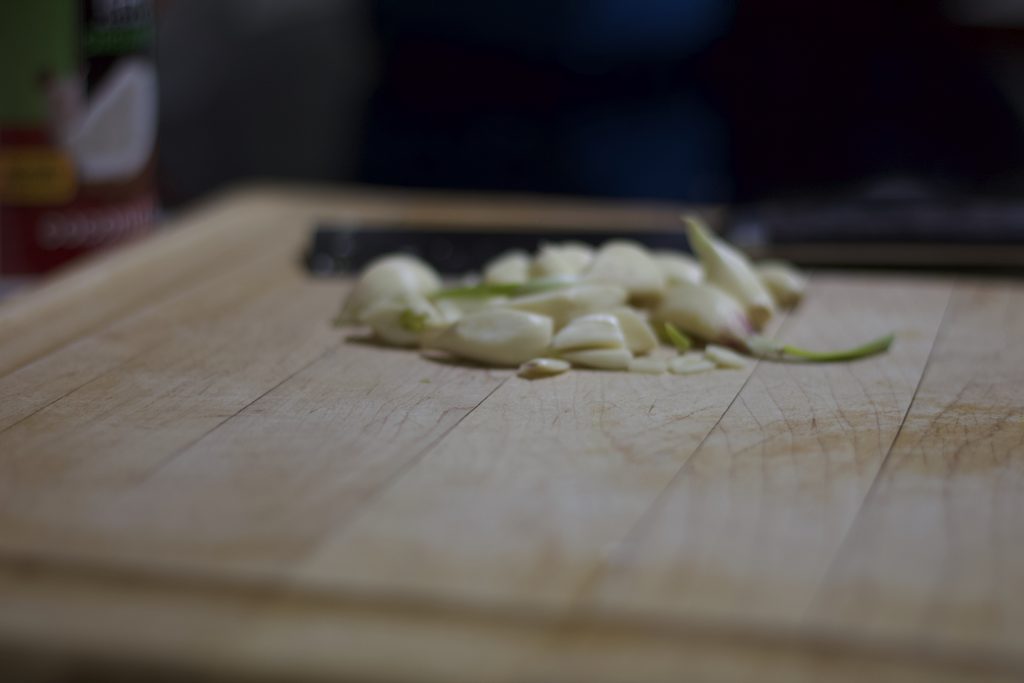 Relatively low angle photo of a wooden cutting board with some garlic further back.