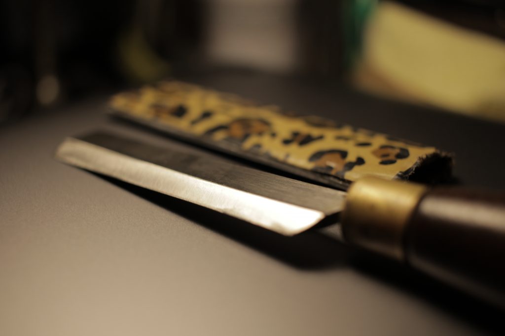 Beveled knife and leopard pattern duct-tape sheath behind it, on top of a brushed aluminum plate.