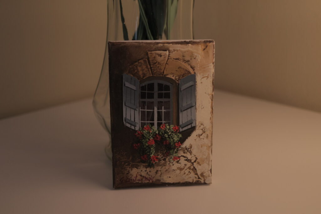 Picture of a painting of a window and flowers on a windowsill against a vase.