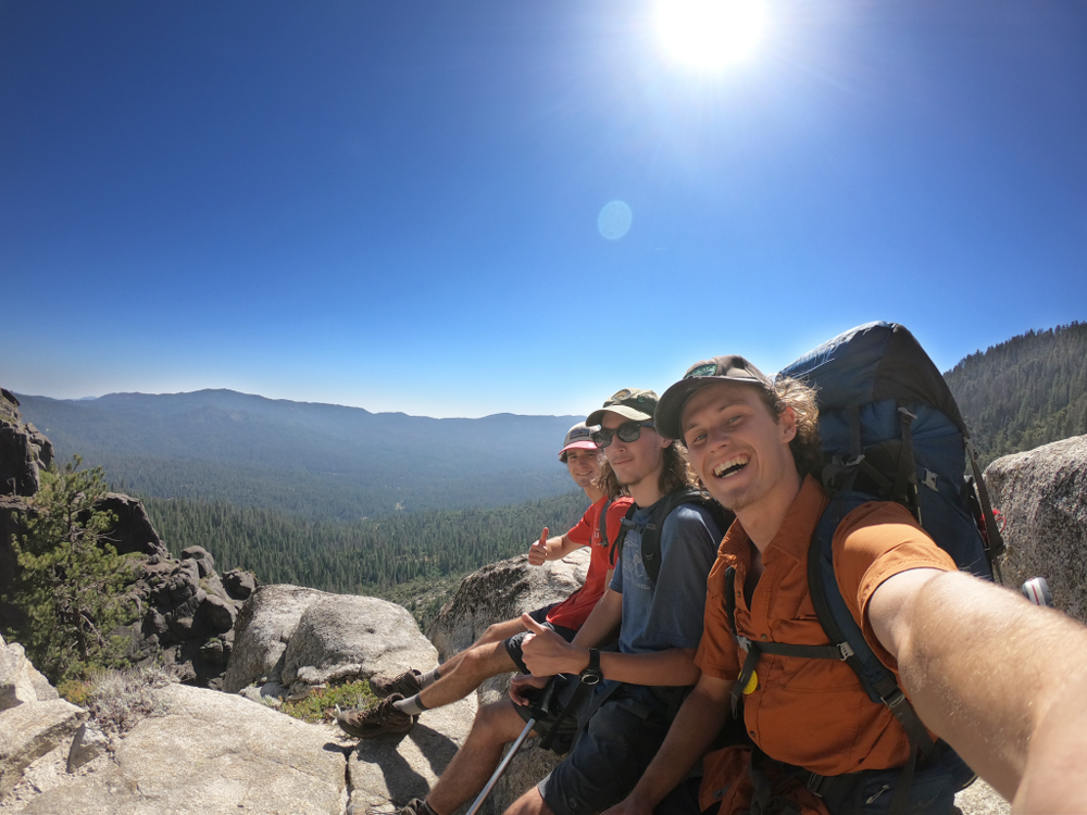 Three backpackers sitting on a rock smiling with a scenic skyline in the background.