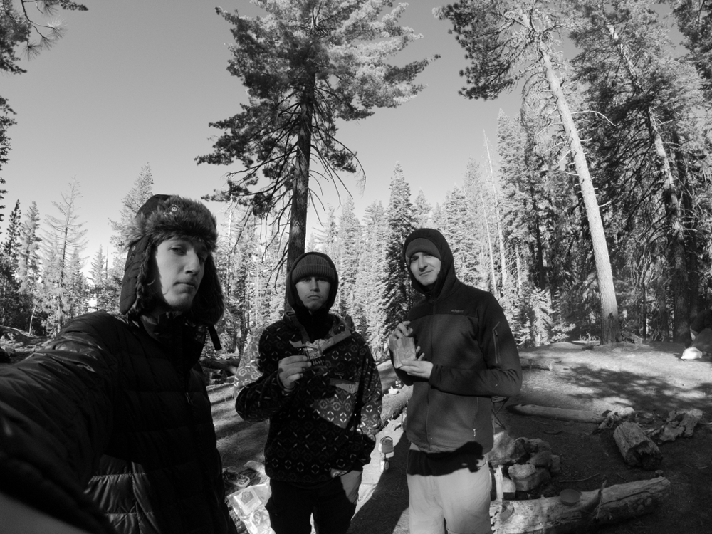Three young men stand in jackets and warm hats snacking on oatmeal and breakfast bars with trees in the background