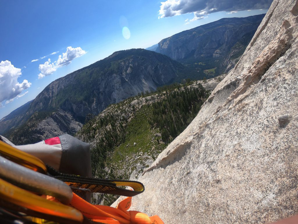 A chalk bag and some carabiners dangle over a steep rock face with small bushes at the bottom.