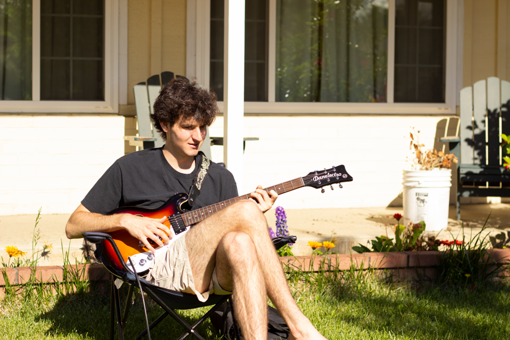 A college student sitting in someone's front yard in a camping chair with a guitar