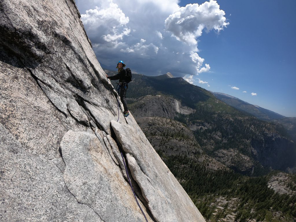 A rock climber with a blue helmet hanging off a rock with trees and mountains far off in the distance.