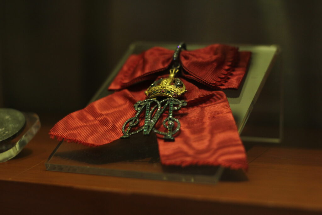 A scarlet red bow brooch with a metal charm attached to it. The charm is a golden crown with little green and red gems with an embroidered letter M underneath it.