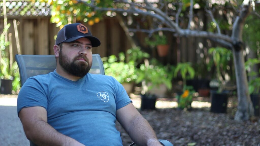 David Miller sitting in his backyard, wearing a hat advertising his woodworking business