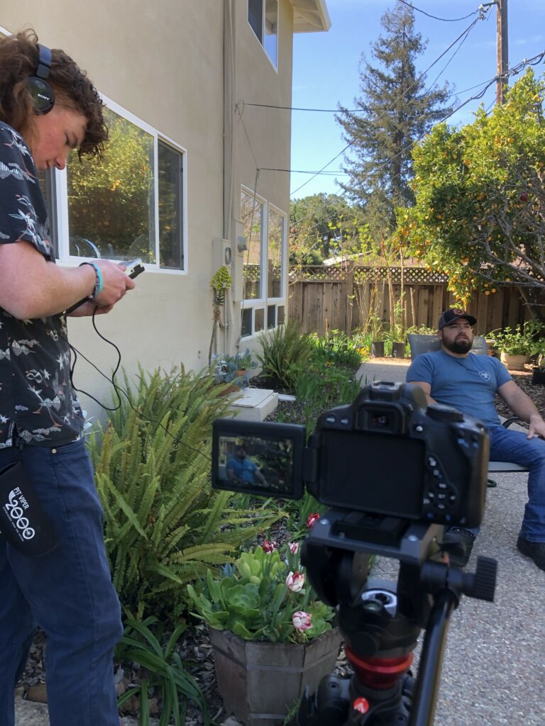 Jack Dees checks the volume of the audio recorder in the backyard of David Miller(the subject of the documentary), as David sits in the background, preparing for his interview. The camera is framed in a close up.