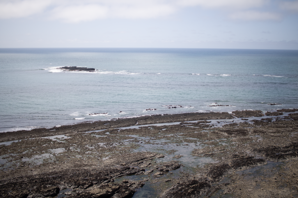 The ocean waving onto a rocky black shore. There are some smaller rocks jutting out of the water further out.
