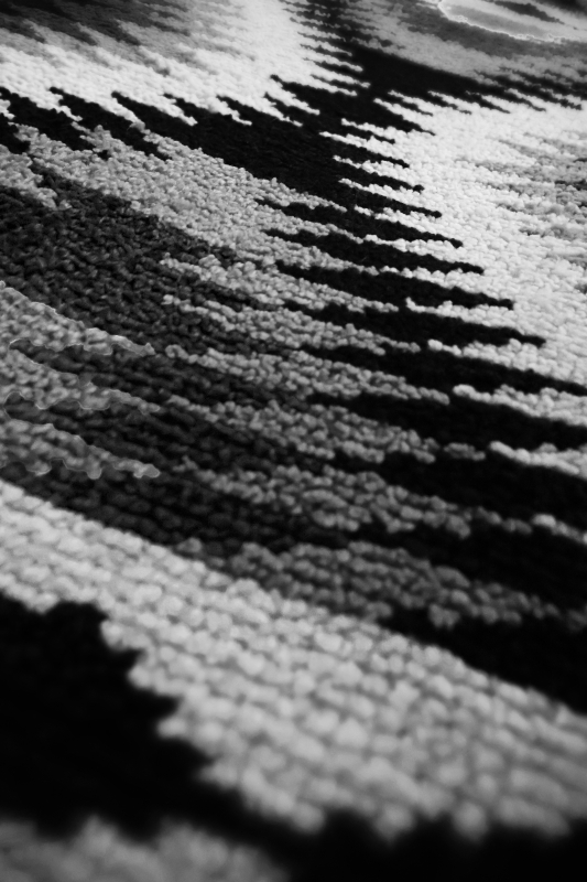 A black and white, portrait photo of a rug taken at an angle. The rug features symmetrical, geometric patterns. The image features a lot of contrast, and the sections of color are distinct. 