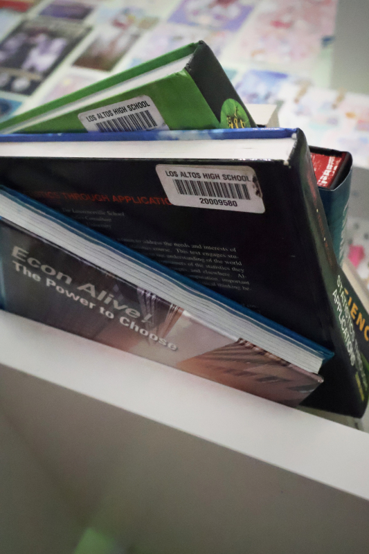 A portrait image taken from a low angle. A stack of school textbooks hangs over the edge of a white table. The background is blurred and shows a mint green wall with many colorful posters