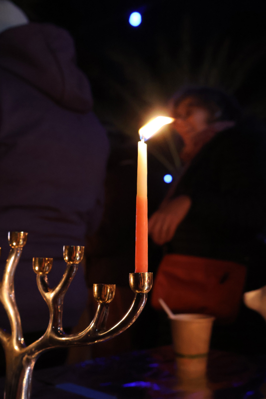 A portrait photo taken outside in the dark. In the center is a lit, orange candle, situated in a chanukiah with its right side visible in half of the frame. The chanukiah is sculpted to look like a tree, with its candle holders being at the tip of each branch. In the background, a woman and a brown paper cup are barely visible.