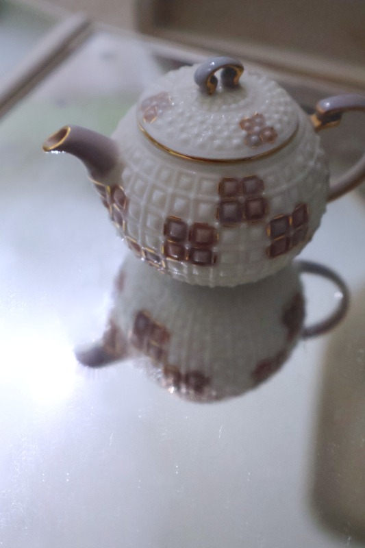 A portrait photo of a small toy teapot. The teapot is ceramic and primarily white. The handles and spout are pale purple with a gold border. The teapot is covered in a square pattern texture, and random sets of four squares are painted pale purple with a gold border on each square. The teapot is in the top of the frame, and it is sitting on a mirror such that its reflection is visible in the bottom of the frame. Positioned near the spout of the reflection is a large bright spot created by the reflection of a lightbulb.