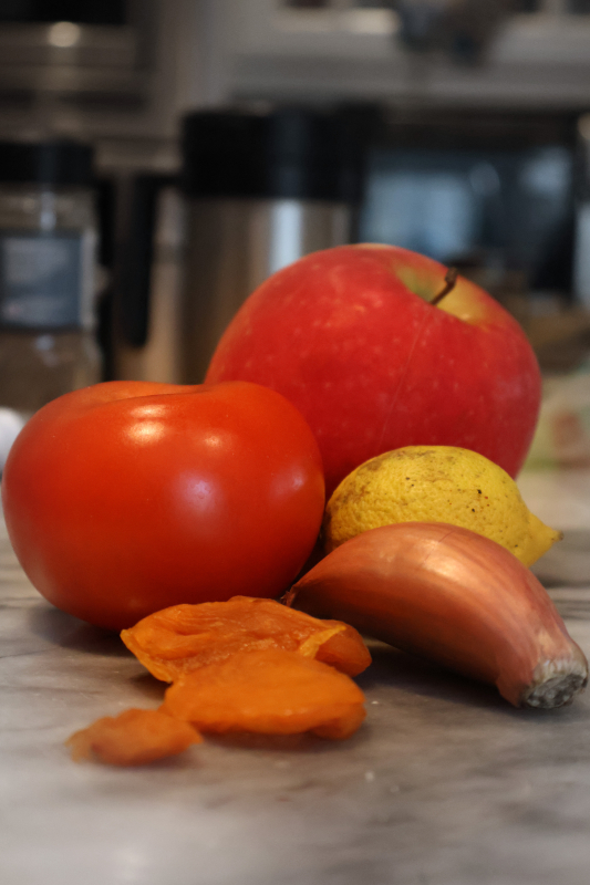 A portrait photograph of an apple, tomato, small lemon, garlic clove, and 3 apricots arranged on a marble countertop in a kitchen. The items feature very warm colors