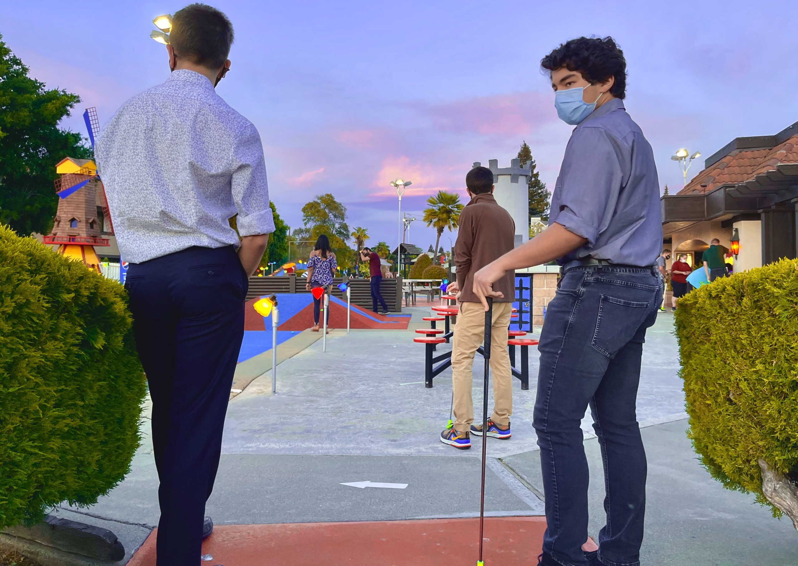 It is dusk at a minigolf place and a few people stand in the foreground looking in various directions. 