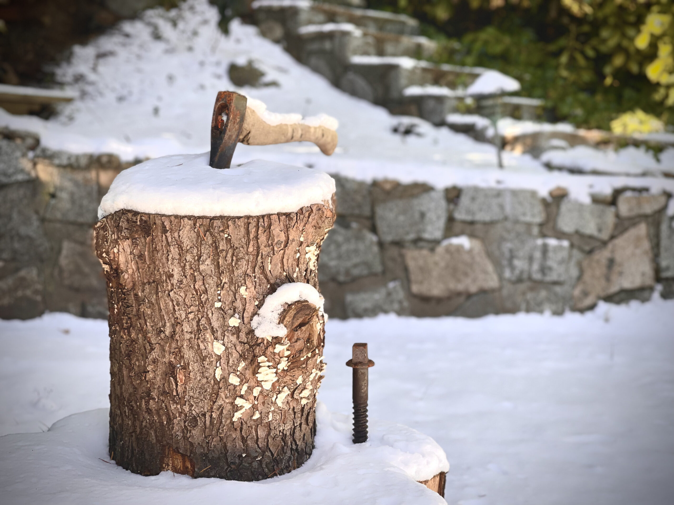 An old axe stuck in a log in front of s tone backdrop and staircase. Snow sits on top of it all. 