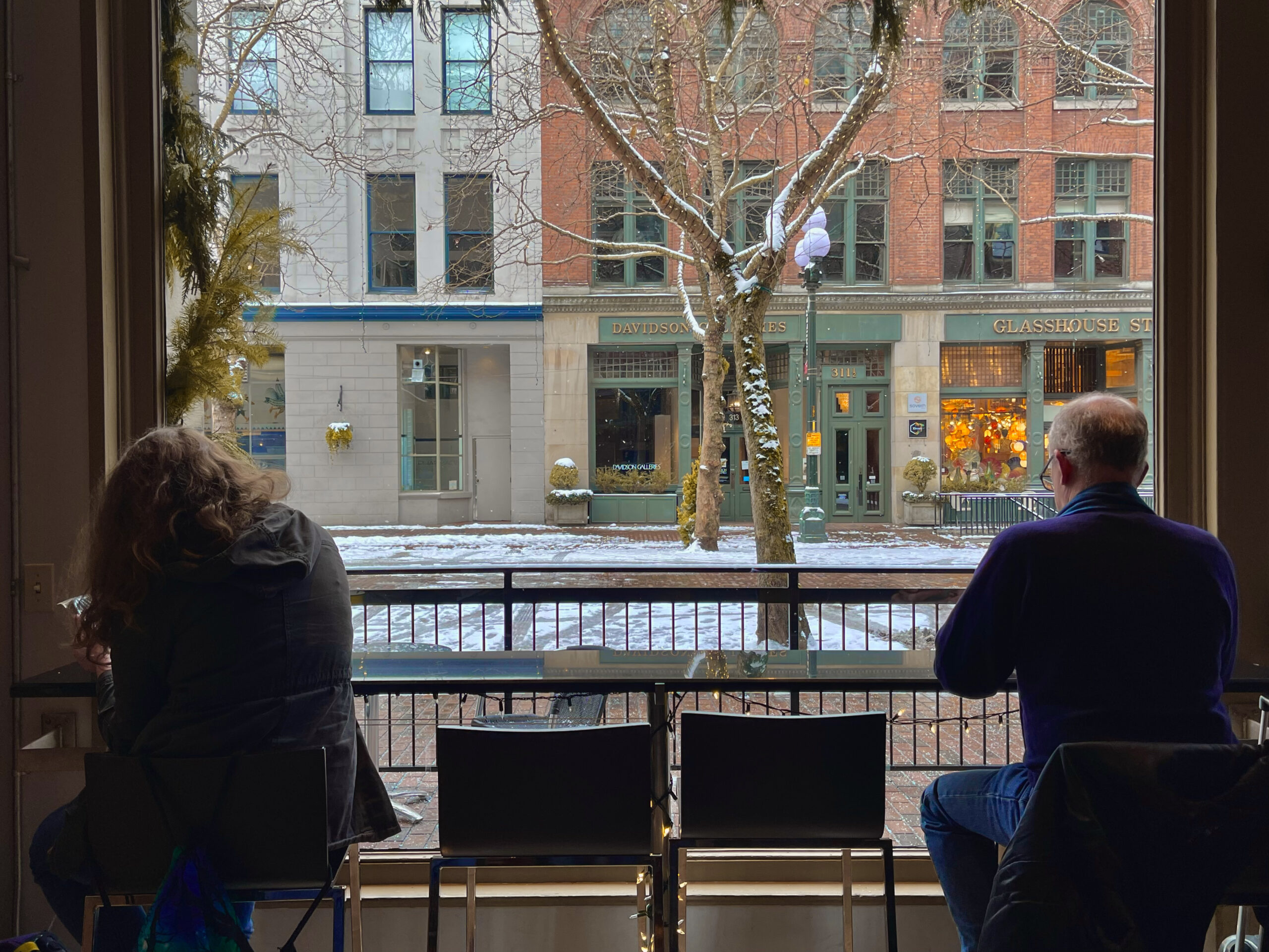 Two people sit in the window of a cafe contrasted against the light of the snowy outside town. 