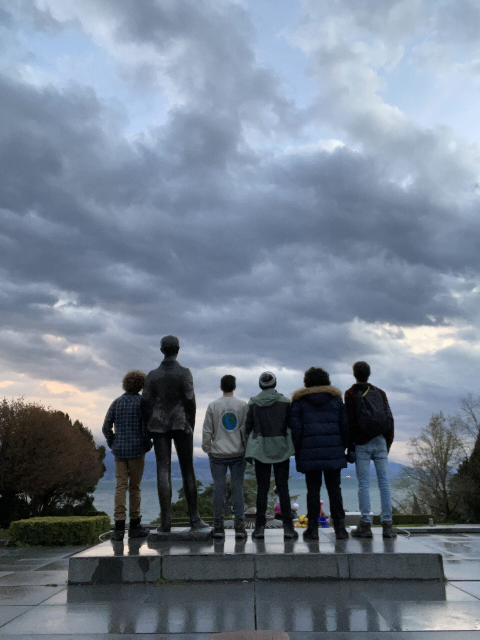 5 boys stare out onto the sunsetting horizon next to an old statue. 