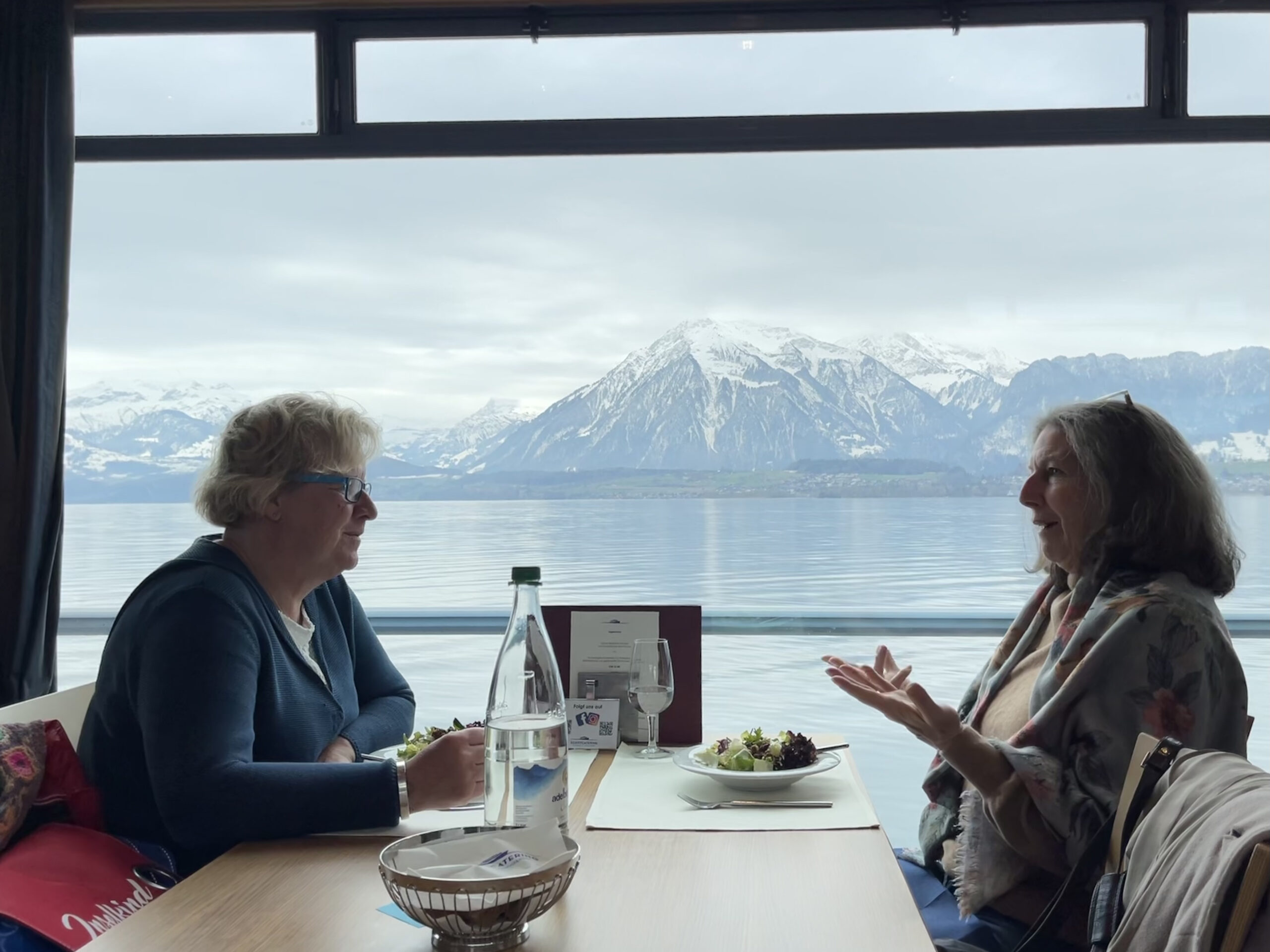 Two ladies sit, eating and talking at a table on a boat with a stark mountain behind them. 