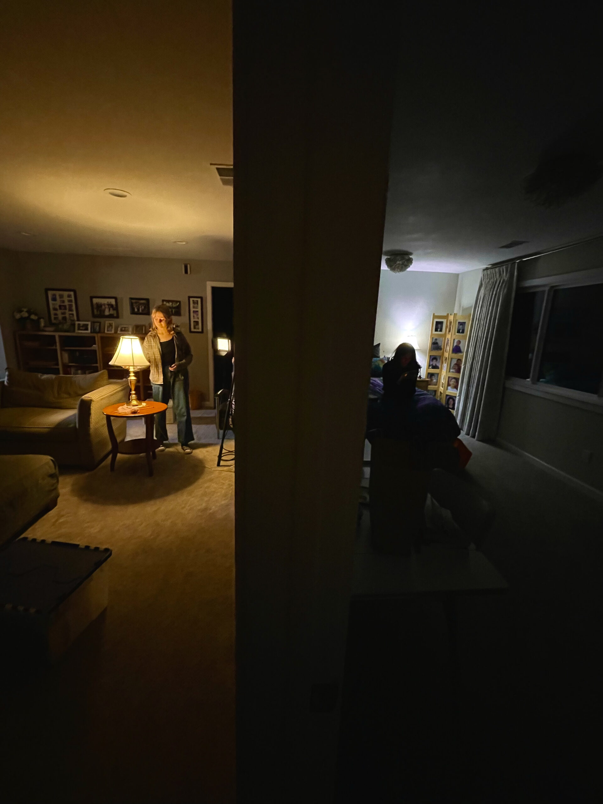 One girl sits in  on a bed in cool darkness while another, on the other side of a wall, turns on a lamp.
