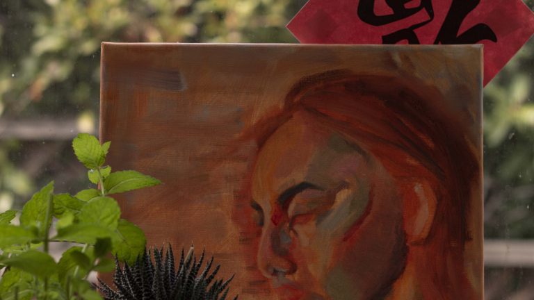 Photo of my culture, depicting a unfinished self-portrait with warm, color-blocked hues. Plants, oil paint tubes, a gold piggy bank, and a ceramic rose frame the background. A Chinese "fu" and bushes make up the background.