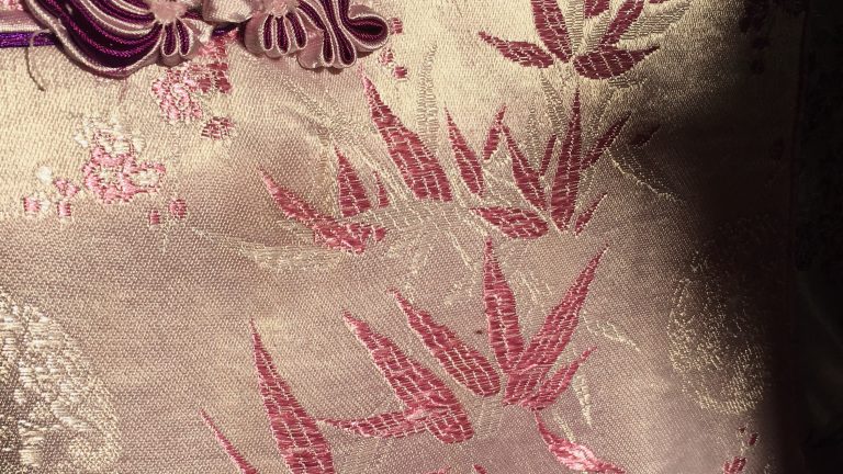 A photo of the details of a traditional Chinese silk dress, chi pao. It’s purple silk fabric in the sun with intricate details.