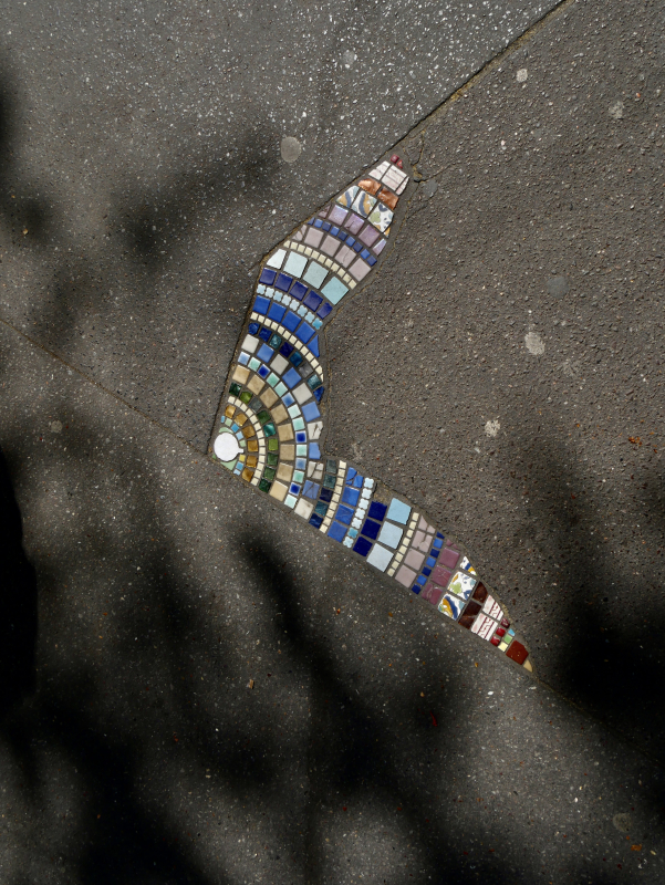 A top-down photo of a cracked sidewalk where the gap has been filled with a colorful mosaic of small ceramic tiles. The mosaic follows the irregular V-shape of the crack, using tiles in shades of blue, teal, green, tan, mauve, and red arranged in concentric arc patterns. The surrounding concrete is grey and textured. Diagonal shadows from a fence or railing fall across the lower portion of the frame.