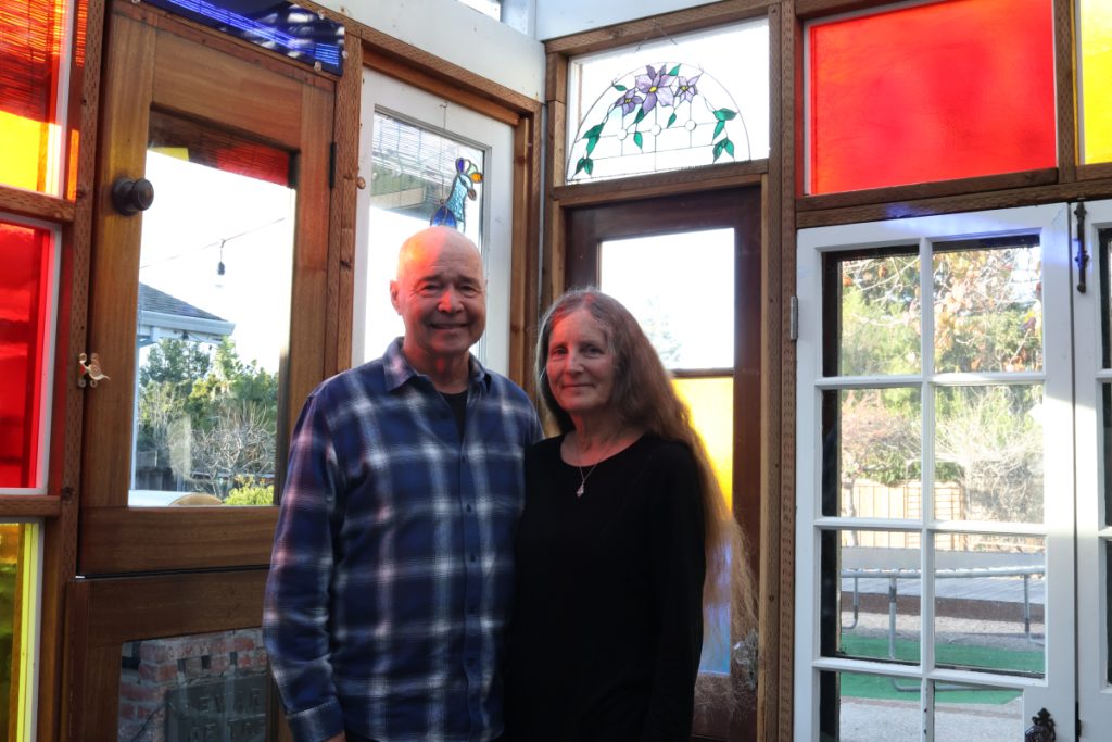 A portrait of two people standing together and smiling inside a room surrounded by stained glass windows and panels. On the left is George Whitehill, an older man with a shaved head wearing a blue plaid flannel shirt. On the right is Jay Whitehill, a woman with long reddish-brown hair wearing a black top and a small pendant necklace. Behind them are wooden-framed doors and windows with colorful stained glass panels in red, yellow, and blue, along with a decorative floral stained glass transom window above the door. Natural light filters in from outside through the clear windows.