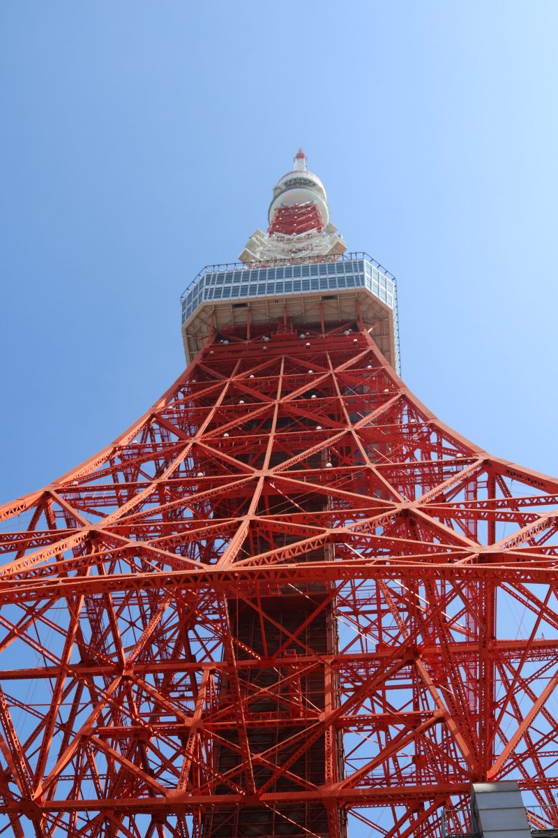 A low-angle, upward-looking photo of Tokyo Tower taken from directly below. The tower's bright red steel lattice framework dominates the frame, with diagonal crossbeams creating a dense geometric pattern that narrows toward the top. An observation deck and the tower's white-and-red upper section are visible near the top of the image. The background is a clear, cloudless blue sky.
