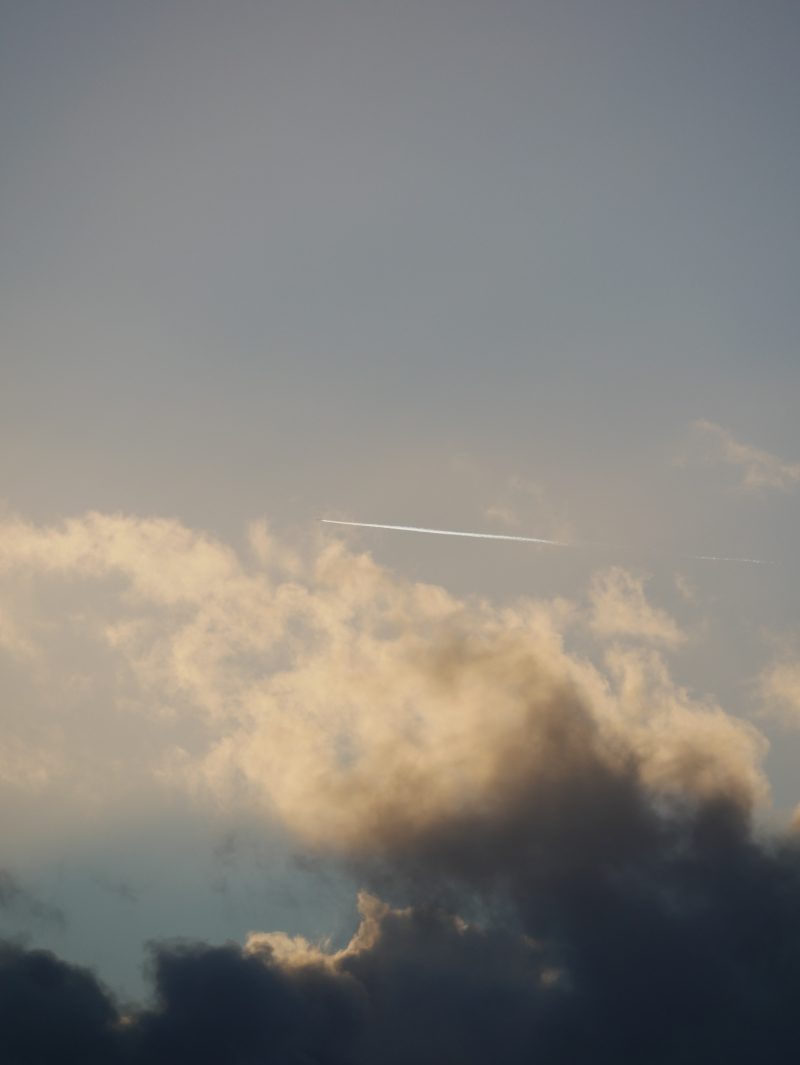 A photo of a dramatic cloudy sky at golden hour. Large storm clouds fill the lower half of the frame, their tops lit in warm golden and amber tones while the bases are dark grey and navy. A thin white airplane contrail cuts diagonally across the middle of the image against a muted blue-grey upper sky. A second faint contrail is visible in the distance to the right.