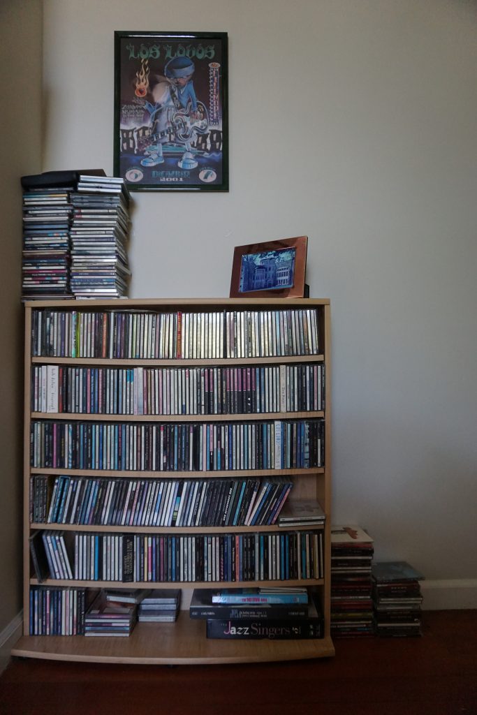 A pciture of what could be a bookshelf but instead of books is filled with CDs. There are stacks of CDs on top of the bookshelf and beside it. On the wall a Los Lobos poster is centered. 