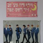 There are two posters of BTS in the picture. One of them is a more traditional postr of the group and the other is a pink concert sign with a Korean phrase on it.