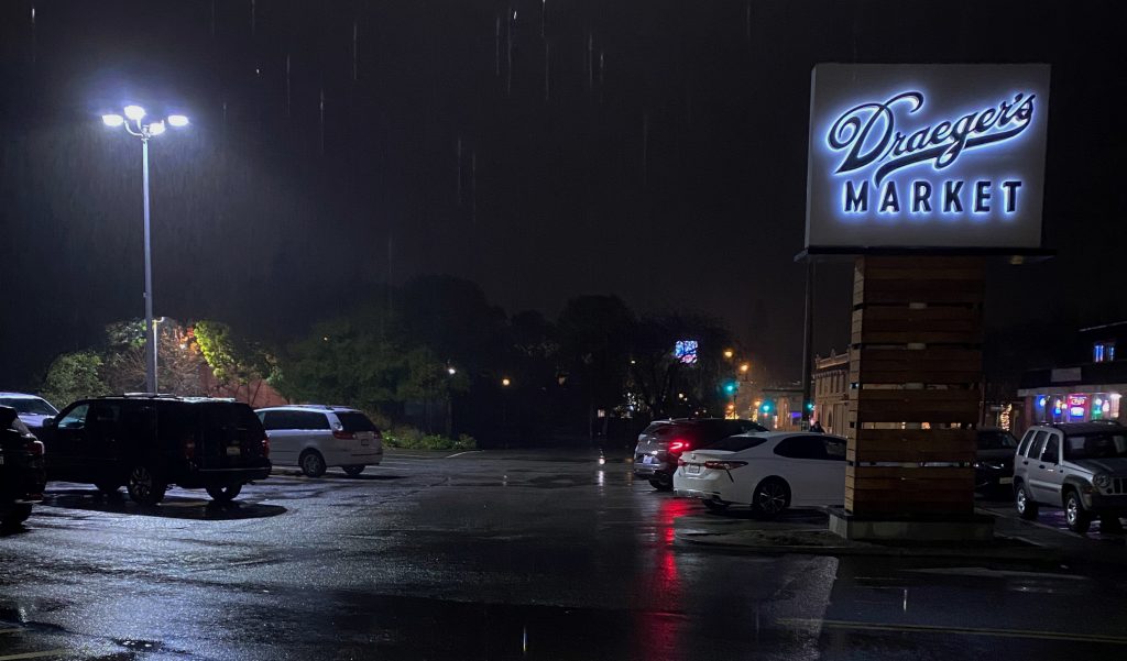 A rainy parking lot is pictured, the sky is dark and it's raining. The sole light in the photo comes from one large overhead light. There is a light up sign saying "Draegers Market" in the right hand corner. 