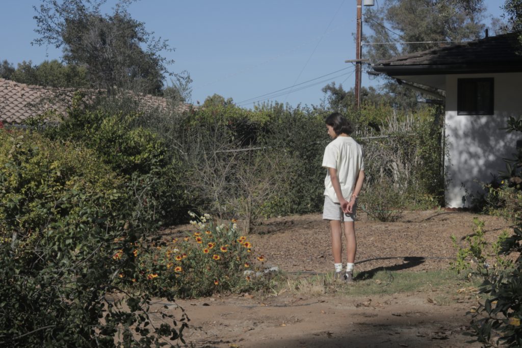 Boy facing away from camera centered using the rule of thirds. Surrounded by a garden and a house in the background.