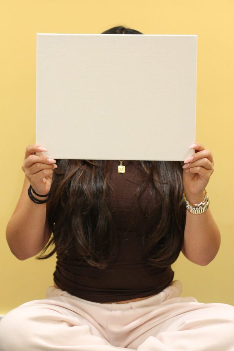 Photo of Mira sitting cross-legged, holding up blank canvas in front of her face