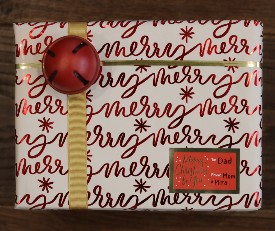 Christmas present wrapped in "merry" wrapping paper with red bell, gold ribbons, and sticker tag.