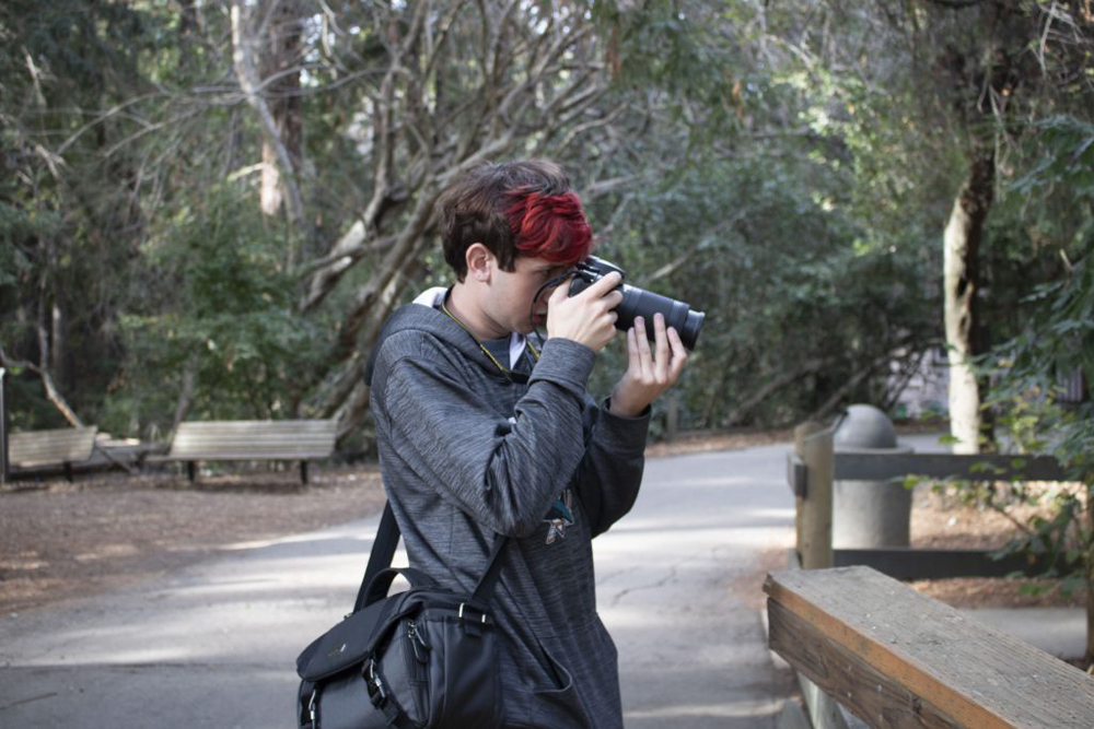 A teenage boy in a grey hoodie stands, facing the right side of the photo (in short, a profile). With partial red-dyed hair (the rest being brown), he stares through the viewfinder of his camera. He is surrounded by forest.