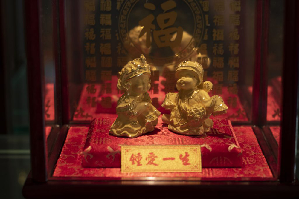 Two golden children in ornamental dress are poised on a small, red pedestal. The children are made of unpolished gold—a matte, ochre material; and small details are made in polished gold, which reflects some light. In front of the two children is a golden-glitter plaque, with red chinese characters on it. Behind the two children is a mirror embedded with golden chinese characters, with one large character in the center.