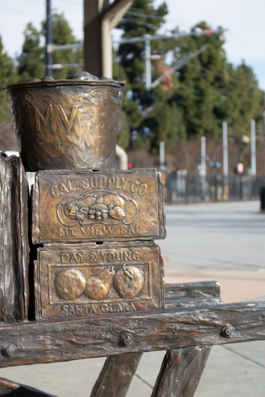 Part of a statue at a train station. A stack 3 high of bronze fruit boxes on the back of a cart are visible, with train tracks in the background.