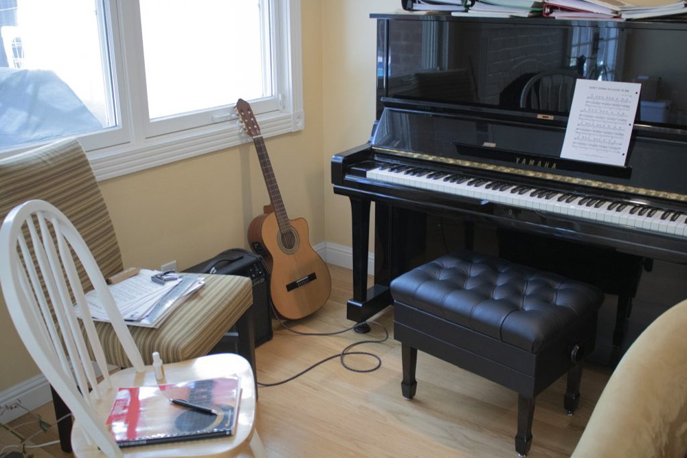 An image of a piano with sheet music on it, a guitar and amp, a book of guitar music, a pen and white-out, a guitar capo. 